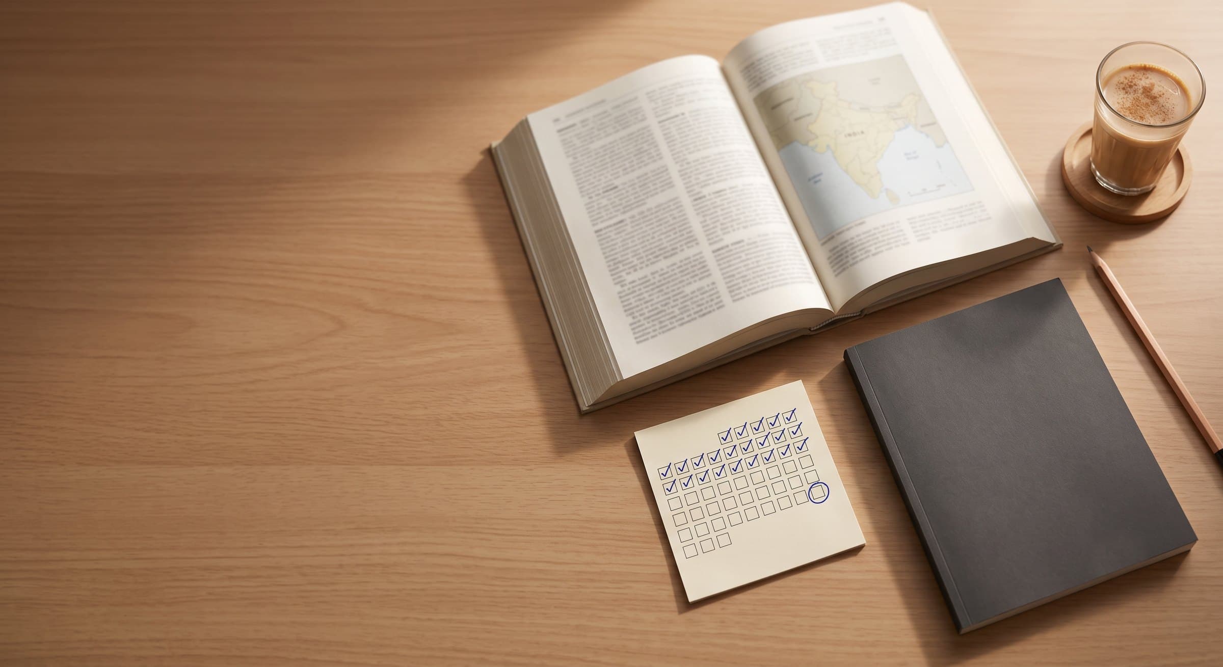 UPSC aspirant's study desk at home: an open reference book with a map of India, a habit tracker filled with blue ticks, a notebook and a glass of chai